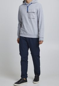 Sweat à capuche gris avec une poche avant et un cordon de serrage bleu, associé à un pantalon bleu marine et des baskets noires. Texture lisse sur toute la surface.