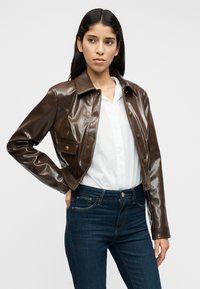 Καφέ δερμάτινο κοντό μπουφάν με γυαλιστερή επιφάνεια, κουμπιά τύπου snap και μία τσέπη στο στήθος. Συνδυάζεται με λευκό πουκάμισο και μπλε τζιν.