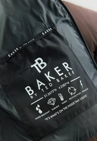 Juodas audinio etiketas su baltais rašmenimis ir simboliais. Įtraukta prekės ženklas "BAKER by TED BAKER" ir priežiūros ikonėlės, nurodančios tokias savybes kaip vandens atsparumas.