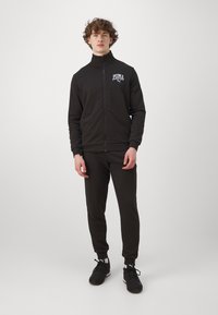 Giacca nera con zip, caratterizzata da un colletto alto e un logo frontale; abbinata a pantaloni jogger neri e scarpe sportive nere.