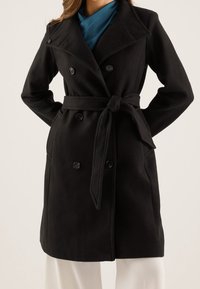 Černý trench coat z vlněné směsi s dvouřadovým zapínáním, širokým límcem a páskem v pase. Má jemnou strukturu a dobře padnoucí střih.