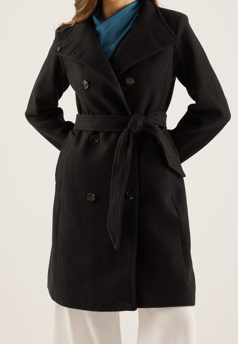 Černý trench coat z vlněné směsi s dvouřadovým zapínáním, širokým límcem a páskem v pase. Má jemnou strukturu a dobře padnoucí střih.