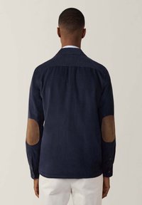 Camicia blu navy con colletto, caratterizzata da toppe in suede marrone sui gomiti e maniche lunghe. Tessuto liscio e vestibilità sagomata.