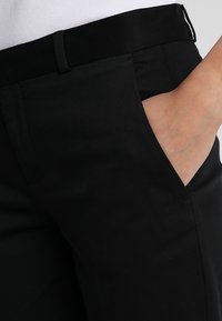 Pantalones negros ajustados con una textura suave, trabillas para cinturón y un bolsillo lateral con un corte relajado. La tela parece ligera y suave.