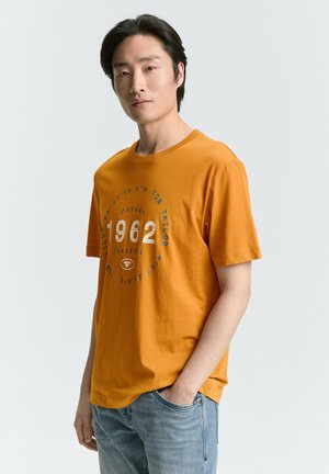 T-shirt in cotone arancione con collo rotondo, maniche corte e una grafica circolare che riporta '1962' e 'Casual Tom Tailor Classics' in blu.
