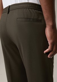 Dunkelolivgrüne Hose mit strukturiertem Stoff, elastischem Bund und einer einzelnen Gesäßtasche auf der rechten Seite.