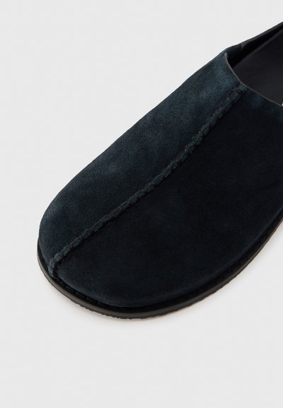 Chausson en daim bleu marine foncé avec une forme arrondie, une couture distincte et une semelle en caoutchouc flexible. Texture lisse et design minimaliste.