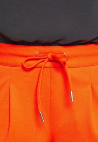 Heldere oranje broek met een trekkoord in de taille, voorzien van zilveren metalen accenten en twee voorkruisen. De stof lijkt glad en textuur te hebben.