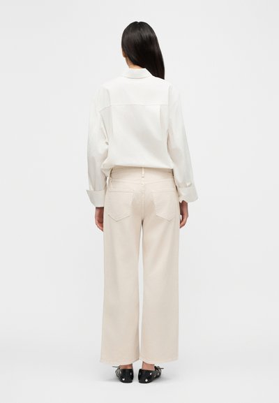 Άνετο λευκό πουκάμισο με μακριά μανίκια, συνδυασμένο με μπεζ παντελόνια φαρδιάς γραμμής. Μαύρα παπούτσια με μεταλλικές λεπτομέρειες ολοκληρώνουν την εμφάνιση.