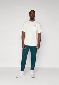 T-shirt de algodão cinza claro com bolso no peito e logótipo, combinada com calças de corrida afuniladas verde-escuro e ténis brancos. Fundo liso.