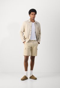 Chemise légère beige, t-shirt blanc, shorts cargo beiges et chaussures à enfiler marron avec des chaussettes blanches, posés devant un fond uni.