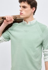 Νεαρός άνδρας με πουλόβερ μέντας και ριγέ πουκάμισο, που κουβαλά καφέ δερμάτινη τσάντα στον ώμο, κοιτάζοντας προς τα πλάγια.