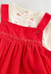 Red corduroy pinafore dress with buttons over white long-sleeve shirt printed with cherries and pink bows.