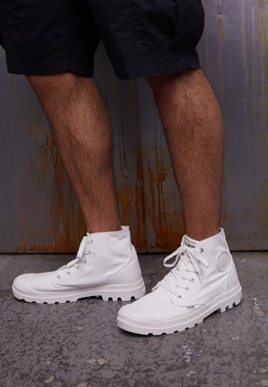 Persona con pantaloncini neri e scarpe da ginnastica bianche alte in piedi su pavimento di cemento testurizzato contro un muro di metallo arrugginito.