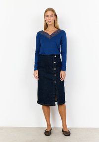 Långärmad blå topp med spetskant, kombinerad med en midiklänning i mörk denim som har en sidoklyvning och knappdetaljer. Svarta nätballerinas.