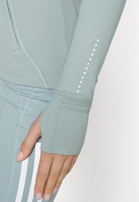 Světlá zelená sportovní bunda s měkkou texturou, kapsou na zip, otvory na palce a jemnými bílými tečkami podél rukávu.