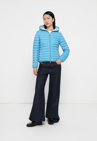 Lichtblauwe pufferjack met horizontale stiksels, een rits aan de voorkant en een capuchon, gecombineerd met donkere wijde jeans en zwarte schoenen.