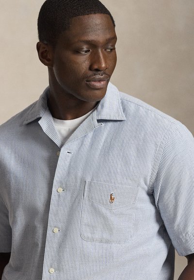 Mladi moški, oblečen v svetlo modro-belo srajco z gumbi in majhnim vezenim logotipom na prsnem žepu, gleda levo.