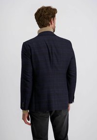 Mann mit lockigem braunem Haar, der ein dunkelkarriertes Blazer über einem beigen Rollkragenpullover trägt, mit dem Rücken zur Kamera vor einem schlichten Hintergrund stehend.