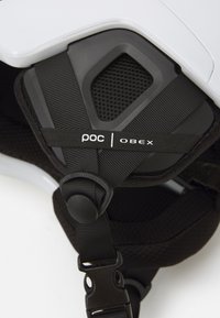 Zbliżenie na kask narciarski POC Obex, z białą powłoką zewnętrzną, czarną siateczkową wyściółką, regulowanymi paskami z elementami metalowymi oraz szczegółem etykiety marki.