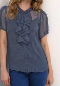 Blå mönstrad blus med volangkrage, korta ärmar och knäppning. Bärs med denimjeans som har en strukturerad yta.