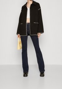 Czarna kurtka z białymi kontrastowymi szwami, z przednimi kieszeniami, połączona z ciemnoniebieskimi dżinsami dzwonami i żółtą torebką. Skórzane botki za kostkę.