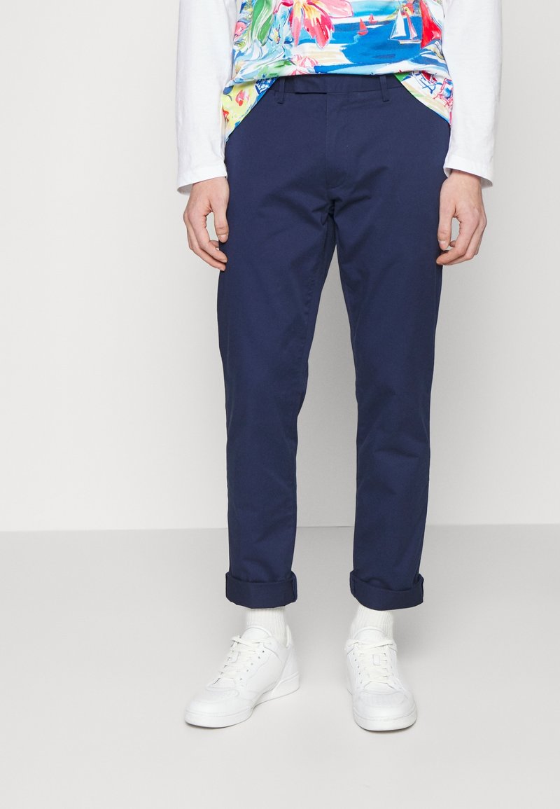 Námořnické kalhoty z bavlny s úzkým střihem, s ohrnutými nohavicemi, nošené s barevnou květinovou košilí a bílými teniskami.