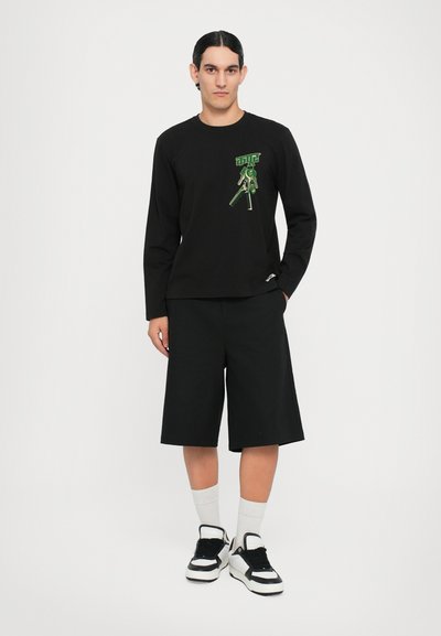 Svart langermet skjorte med grønt grafisk design, kombinert med svarte knelange shorts, hvite sokker og svart-hvite joggesko.