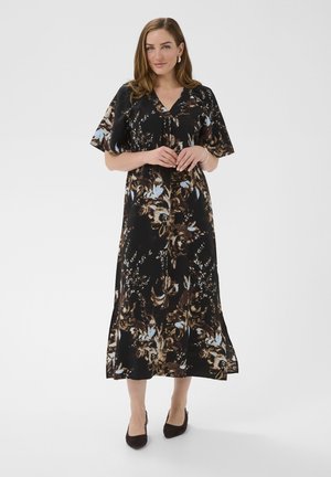 Kobieta w czarnej, kwiecistej sukience midi z krótkim rękawem, stojąca z delikatnie złączonymi dłońmi, w czarnych szpilkach na jednolitym tle.