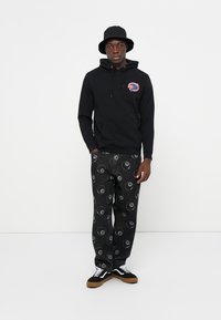Zwarte hoodie met logo, gecombineerd met zwarte broek met een yin-yang patroon. Model draagt een zwarte bucket hat en sneakers met gumzolen.