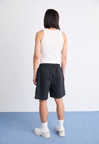 Person with shoulder-length dark hair wearing a white tank top, black shorts, white socks, and white sneakers standing on blue floor against white wall.