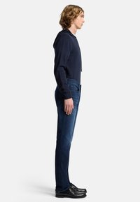 Felpa navy con cappuccio e cordoncino, abbinata a jeans blu scuri e scarpe nere. Il modello sta in posa di lato, mostranndo la vestibilità e i dettagli di design.
