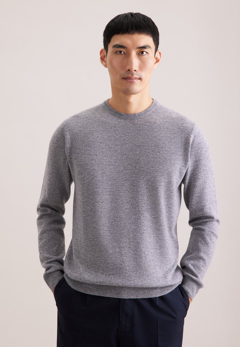 Grauer Strickpullover mit langen Ärmeln, Rundhalsausschnitt und gerippten Bündchen. Einfaches Design mit weicher Textur. Über einer dunklen Hose getragen.