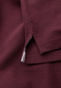 Primer plano del dobladillo de una camiseta polo burdeos con abertura lateral y una pequeña etiqueta de tela blanca visible parcialmente dentro de la abertura.
