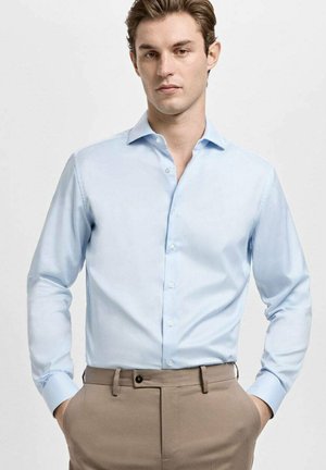 Hellblaues Hemd mit Knöpfen, Kragen, langen Ärmeln und glatter Textur, kombiniert mit beigen Hosen, Hände in den Taschen.