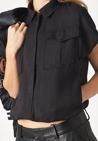 Chemise noire à manches courtes avec boutons et col, dotée de deux poches poitrine et d'un tissu texturé. Coupe ajustée, se termine au-dessus de la taille.