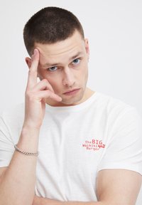 Camiseta de algodón blanca con texto gráfico rojo "The BIG Waikiki Grill Burger" en el pecho. El modelo tiene el cabello corto y una pulsera de plata.
