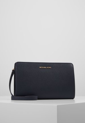 Czarna damska portmonetka Michael Kors z logo w kolorze złota i teksturowaną powierzchnią, umieszczona na białym postumencie na tle jasnoszarym.