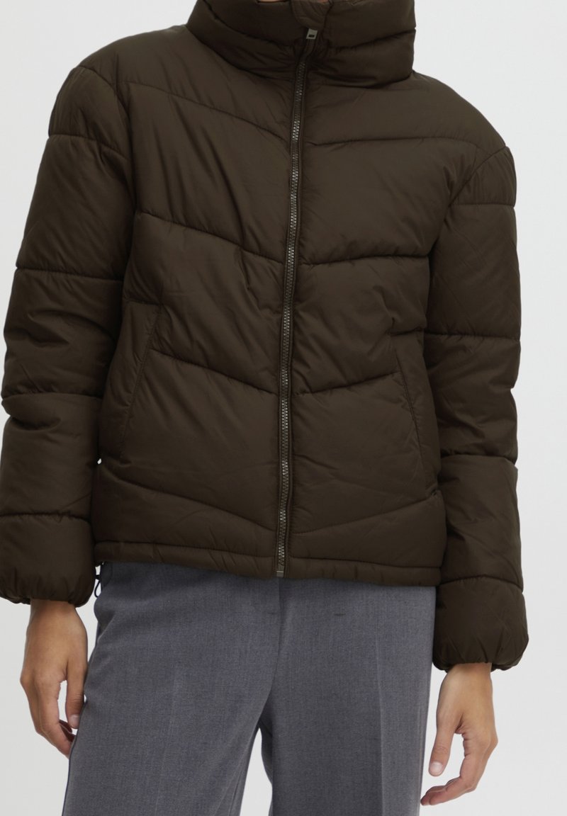 Personne portant une doudoune matelassée marron foncé avec une fermeture éclair à l'avant et un pantalon gris, visible des épaules aux hanches.