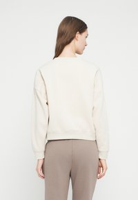 Γυναίκα με την πλάτη στραμμένη, φοράει ένα κρεμ φούτερ και παντελόνια σε απόχρωση ταουπ, πάνω σε απλό λευκό φόντο.