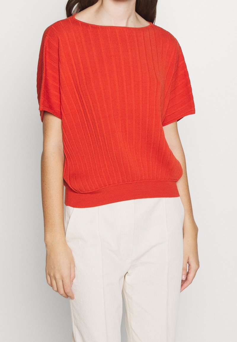 Top de punto rojo acanalado con escote amplio y mangas cortas, que presenta un ajuste relajado y un diseño en color sólido, combinado con pantalones claros.