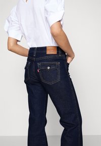 Jean en denim bleu foncé avec une taille haute, des poches arrière et un écusson en cuir. Le tissu présente une texture subtile et des accents de couture.