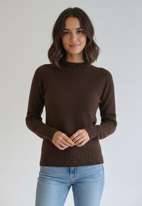 Brązowy sweter z dzianiny z wysokim kołnierzem, teksturowanym wzorem na ramionach i krawędzi oraz prążkowanymi mankietami, noszony na jasnoniebieskich dżinsach.