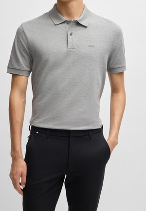 Mann trägt ein hellgraues kurzärmeliges Poloshirt mit „BOSS“-Logo und schwarze Hose, steht vor einfachem Hintergrund.