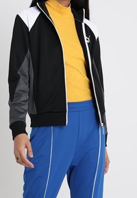 Svart och grå zip-up jacka med vita accenter, tillsammans med en ribbad gul polotröja. Modellen bär blå byxor med vita ränder.