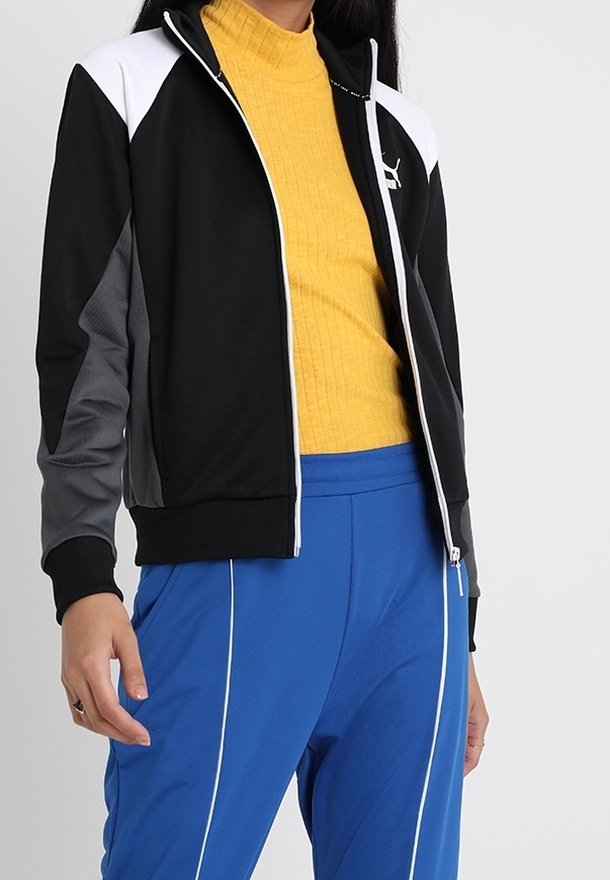 Svart och grå zip-up jacka med vita accenter, tillsammans med en ribbad gul polotröja. Modellen bär blå byxor med vita ränder.