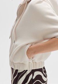 Světle béžová mikina z měkké tkaniny se zipem vpředu, šňůrkou a elastickým lemem. Obsahuje boční kapsu a krátké rukávy.