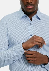 Hellblau gemustertes Hemd mit kleinen marineblauen Punkten, mit Button-Down-Kragen und einzelnen Manschettenknöpfen; der Stoff wirkt glatt und leicht.