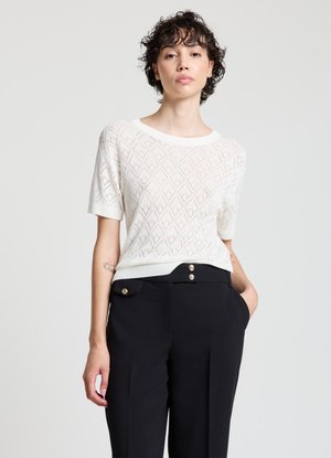 Maglione bianco a maniche corte lavorato a maglia con motivo a rombi, abbinato a pantaloni neri con bottoni dorati e tasche laterali.