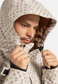 Mężczyzna poprawiający zamek błyskawiczny oraz kaptur beżowej, wzorzystej kurtki pikowanej z brązowymi akcentami logo, pokazanej w zbliżeniu.